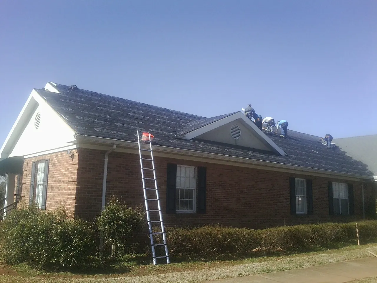 Crew tearing shingles off a brick ranch roof with a ladder set — Metal Roof Repair work by our crew in Mendenhall