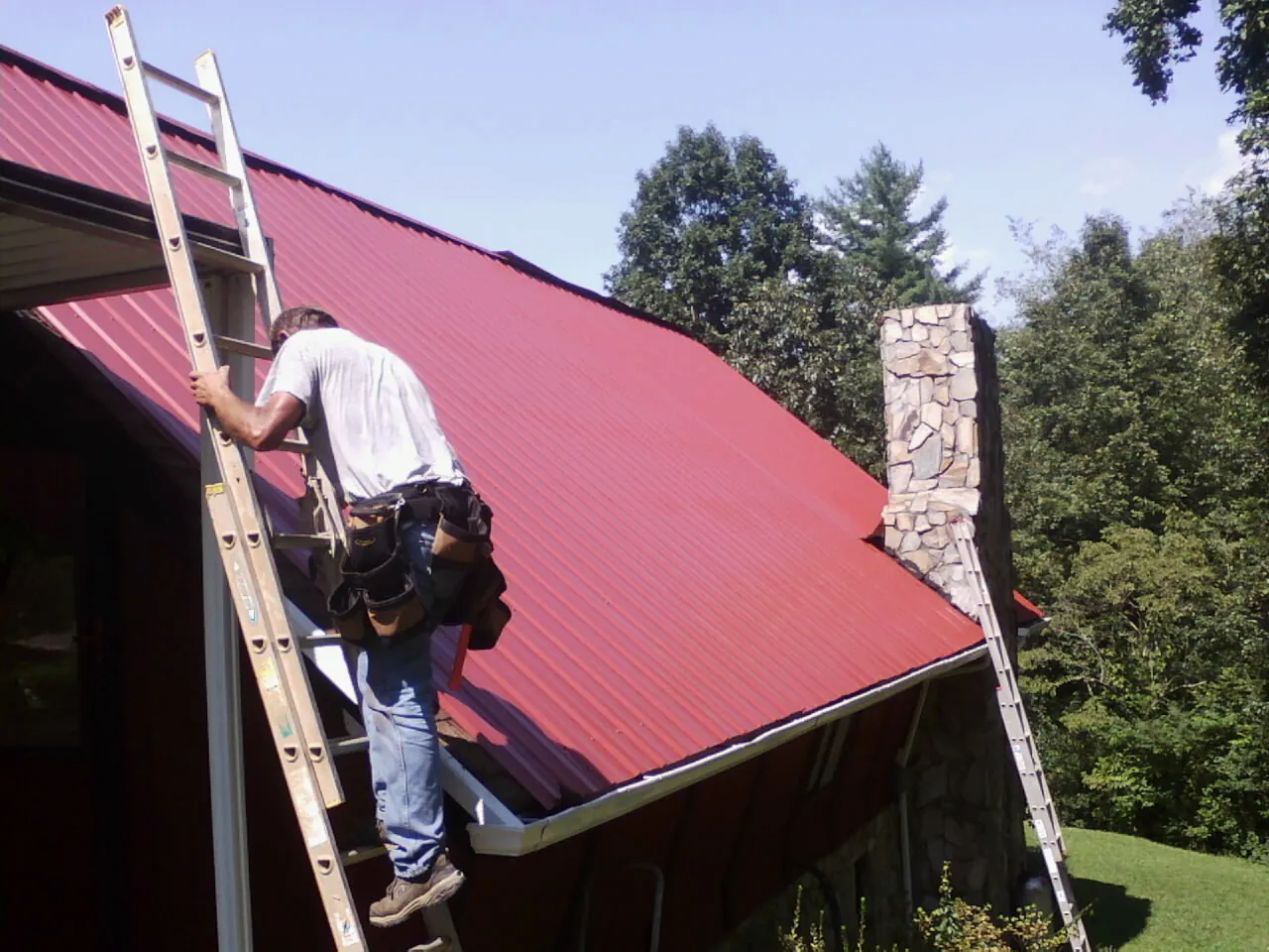 Roofer on a ladder beside a red metal roof and stone chimney — Soffit & Fascia Repair by Roofing Brothers in Mendenhall